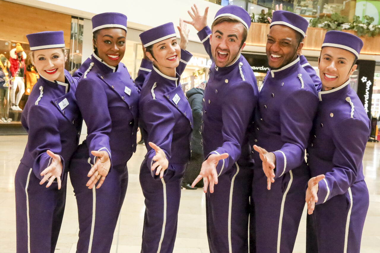 Performers dressed in purple bell hop uniforms smile into the camera