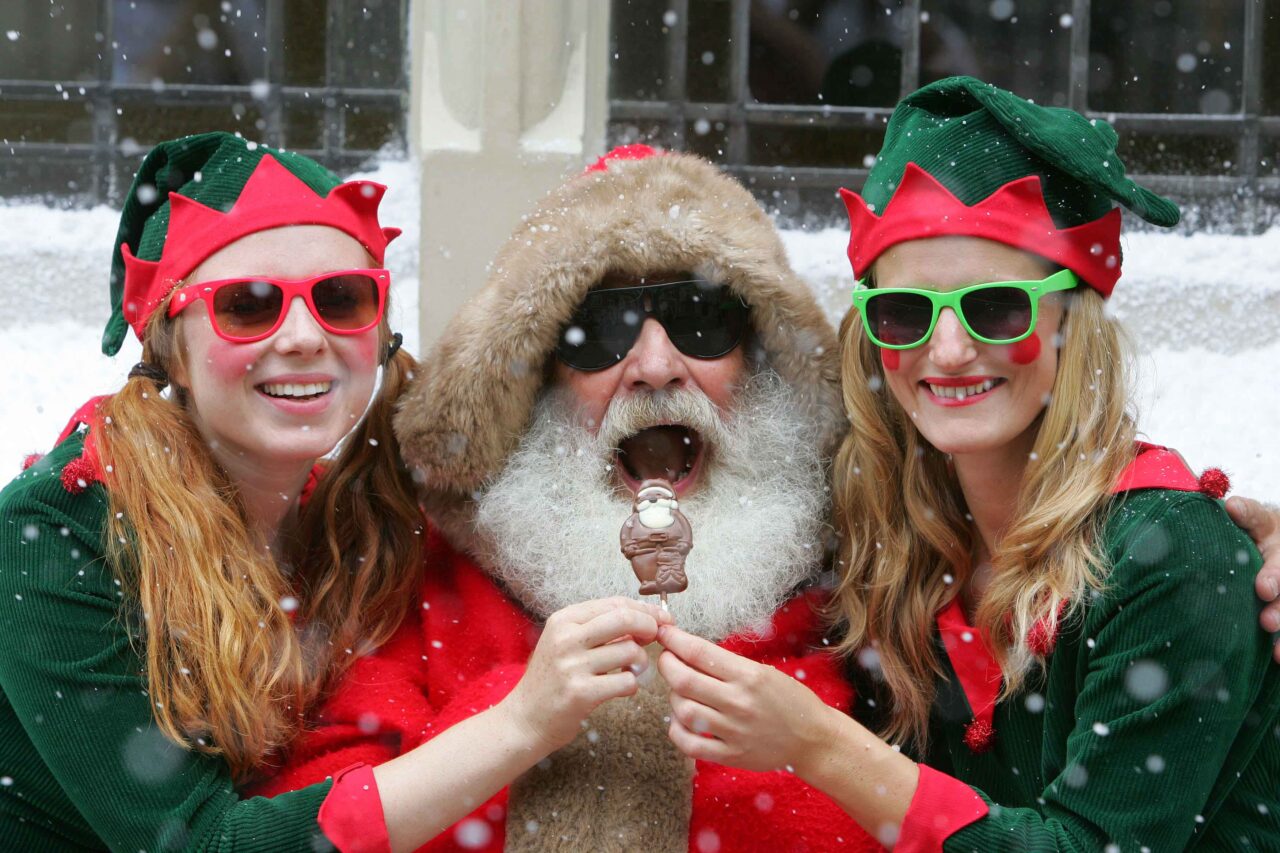 Santa in sunglasses eats a chocolate flanked by two elves