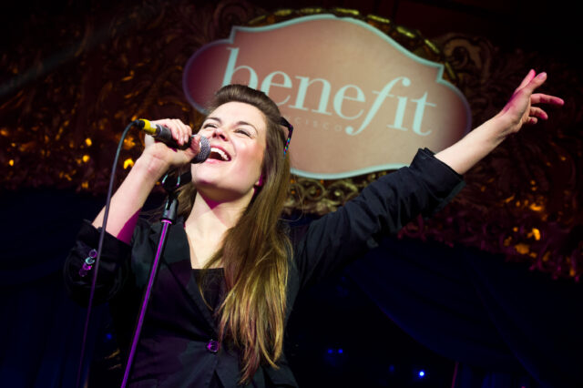 A singer sings into a microphone in front of Benefit branding