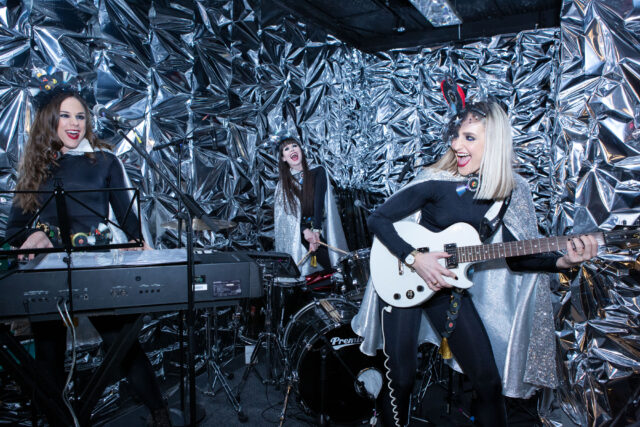 Singers with instruments in front of a shiny silver curtain