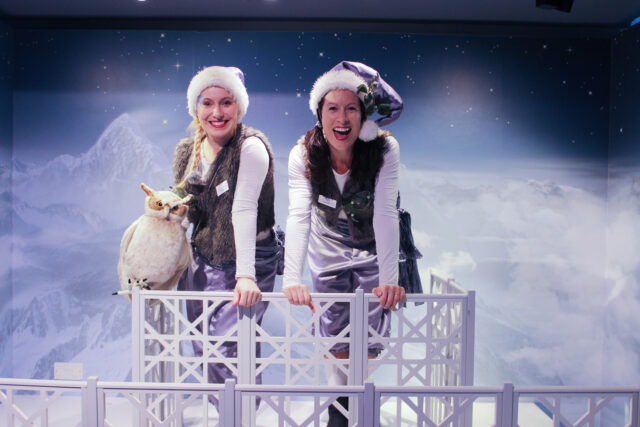 Two elves stand on a white balcony in purple and white hats with a snowy background