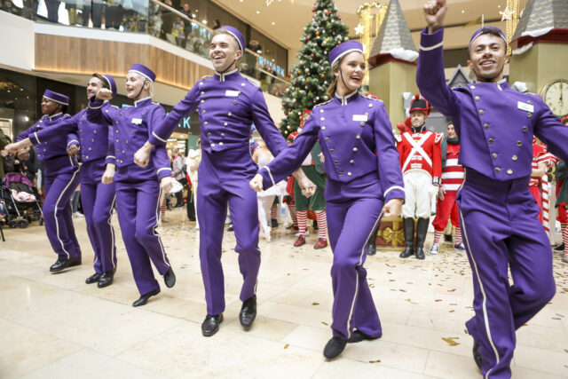 A line of entertainers dressed as purple bell hops dance