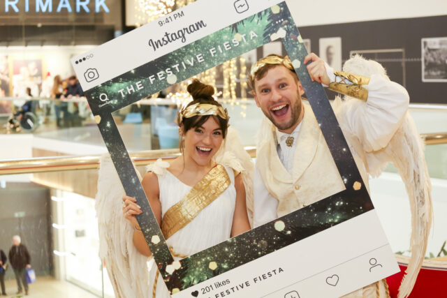 Two people dressed in white and gold hold an Instagram photo frame and smile