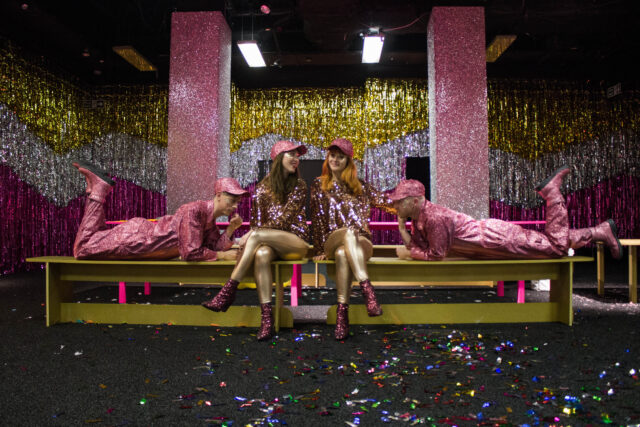 Four people dressed in glitter pose in a room full of glitter decorations