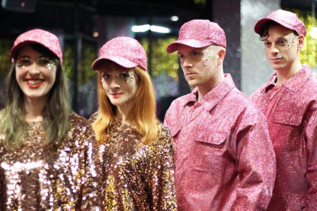 Four performers dressed in pink and gold glitter outfits pose