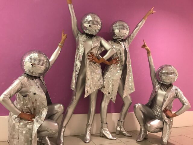 Four people in disco ball costumes pose against a pink wall