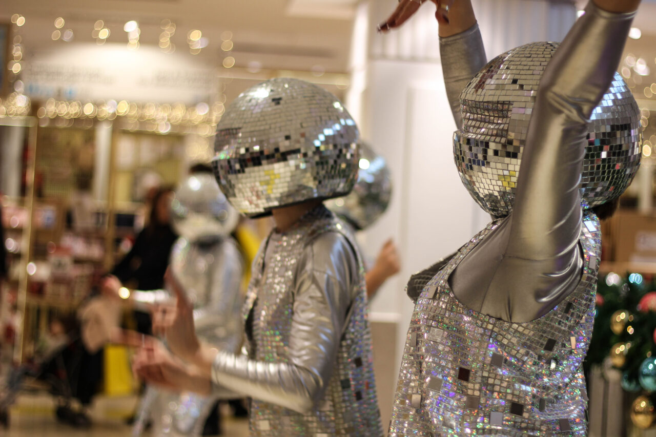 Two people in mirrored outfits with disco ball hats dance